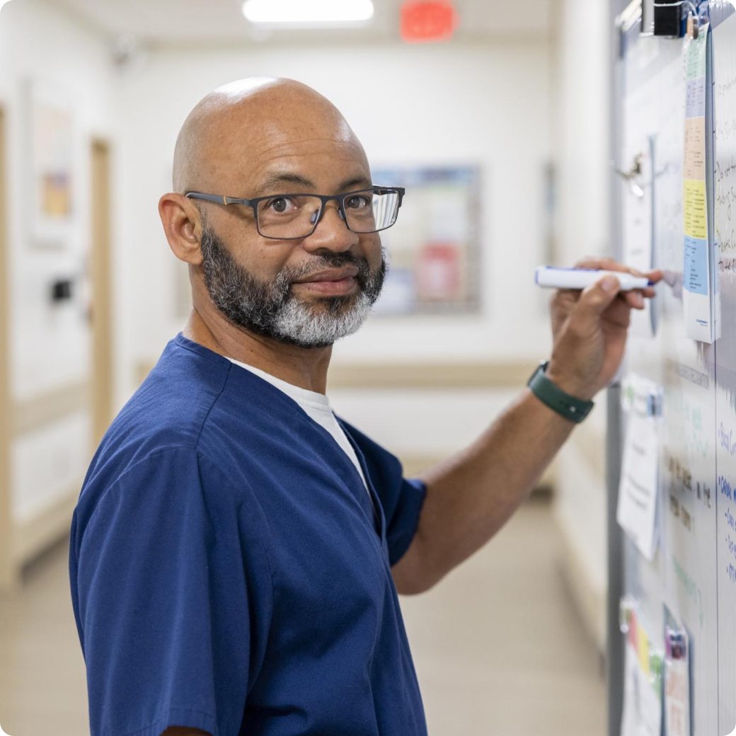 A male nurse in blue scrubs is looking at the camera while writing on a whiteboard.