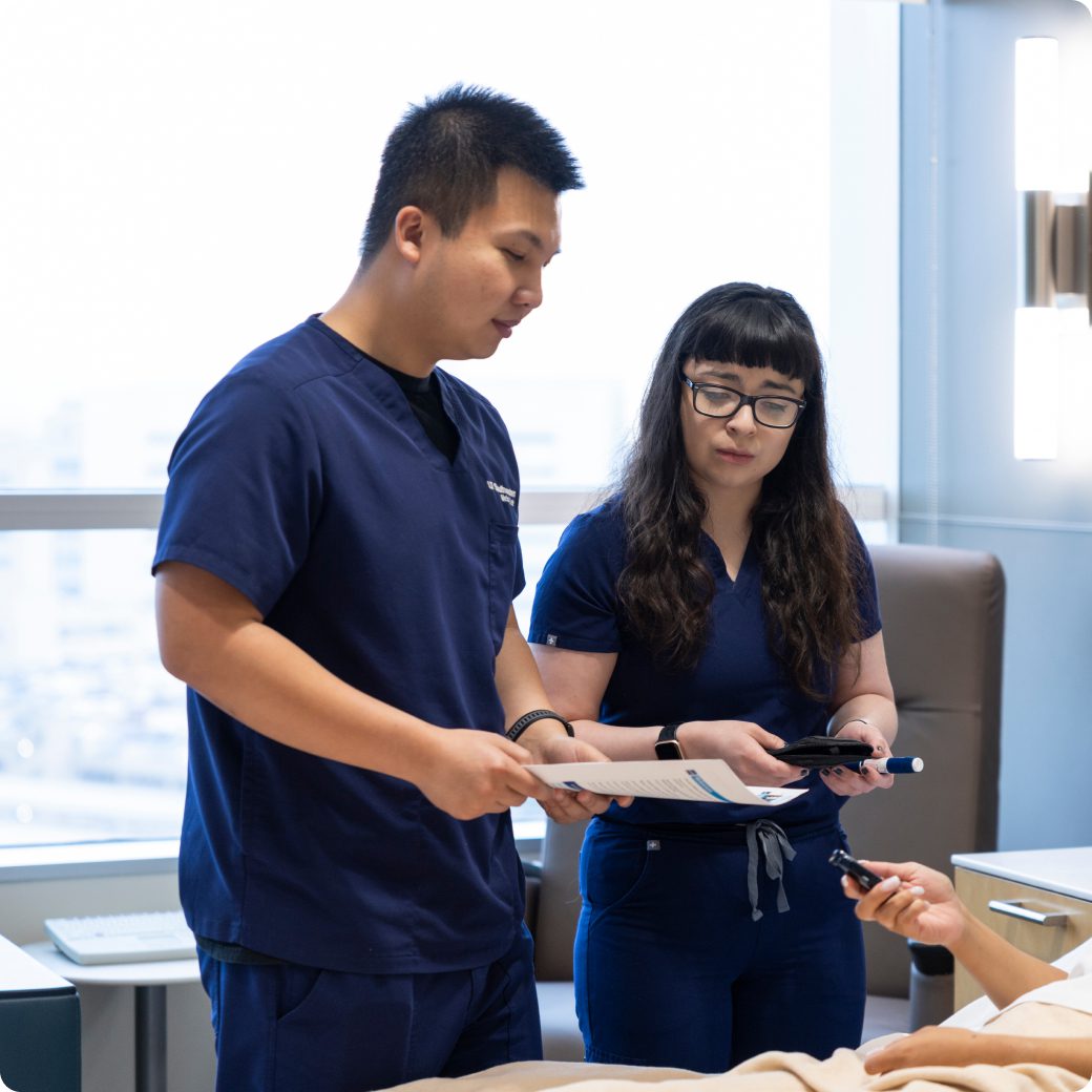 Two hospital nurses in blue scrubs discussing paperwork with a patient in a hospital room.