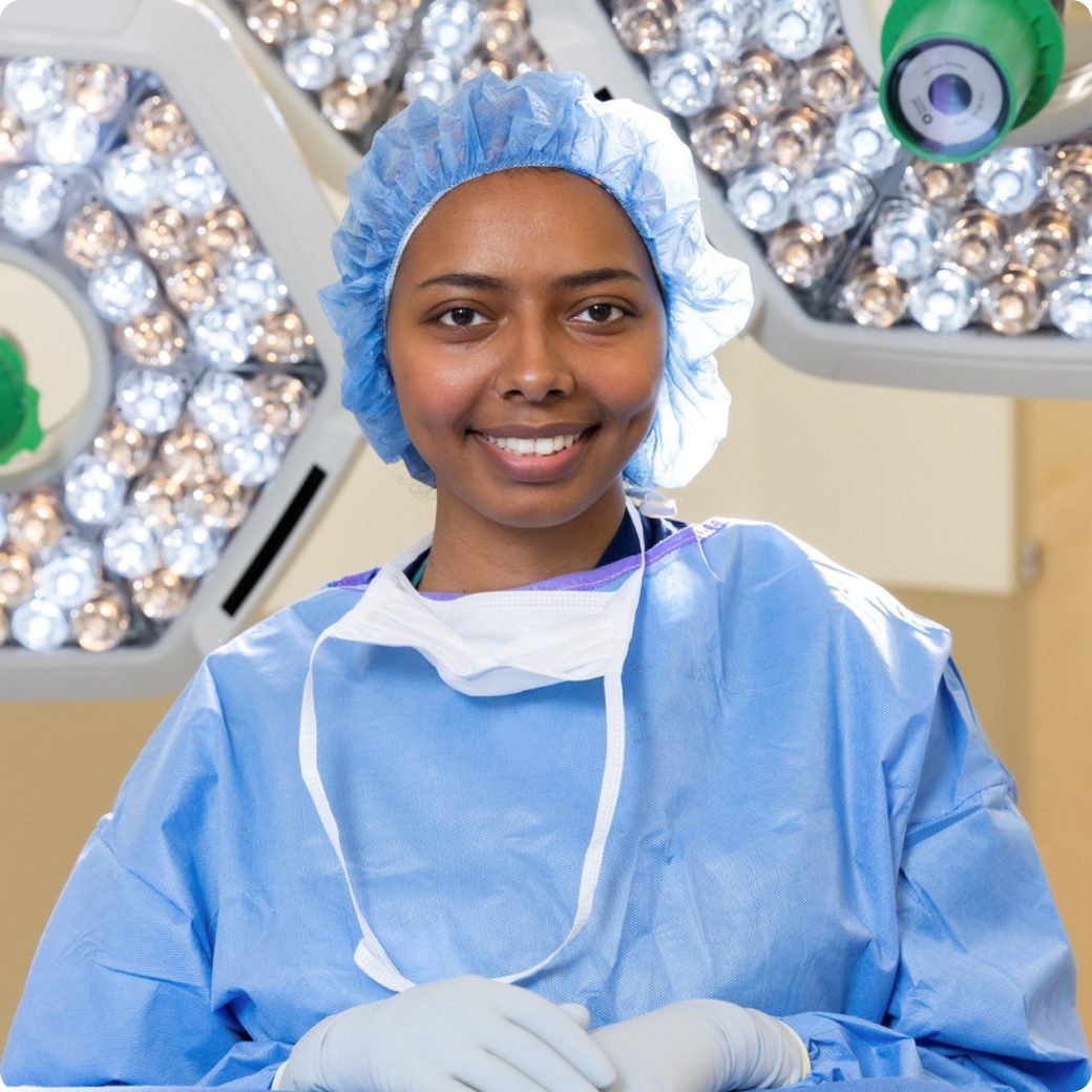A female nurse in a blue surgical gown is looking at the camera, standing in an operating room.