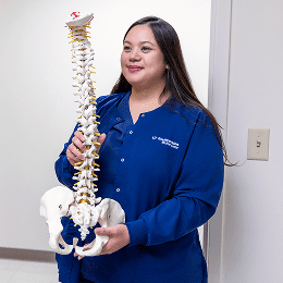 A female nurse in blue medical scrubs holding a life-size model of the human spine.