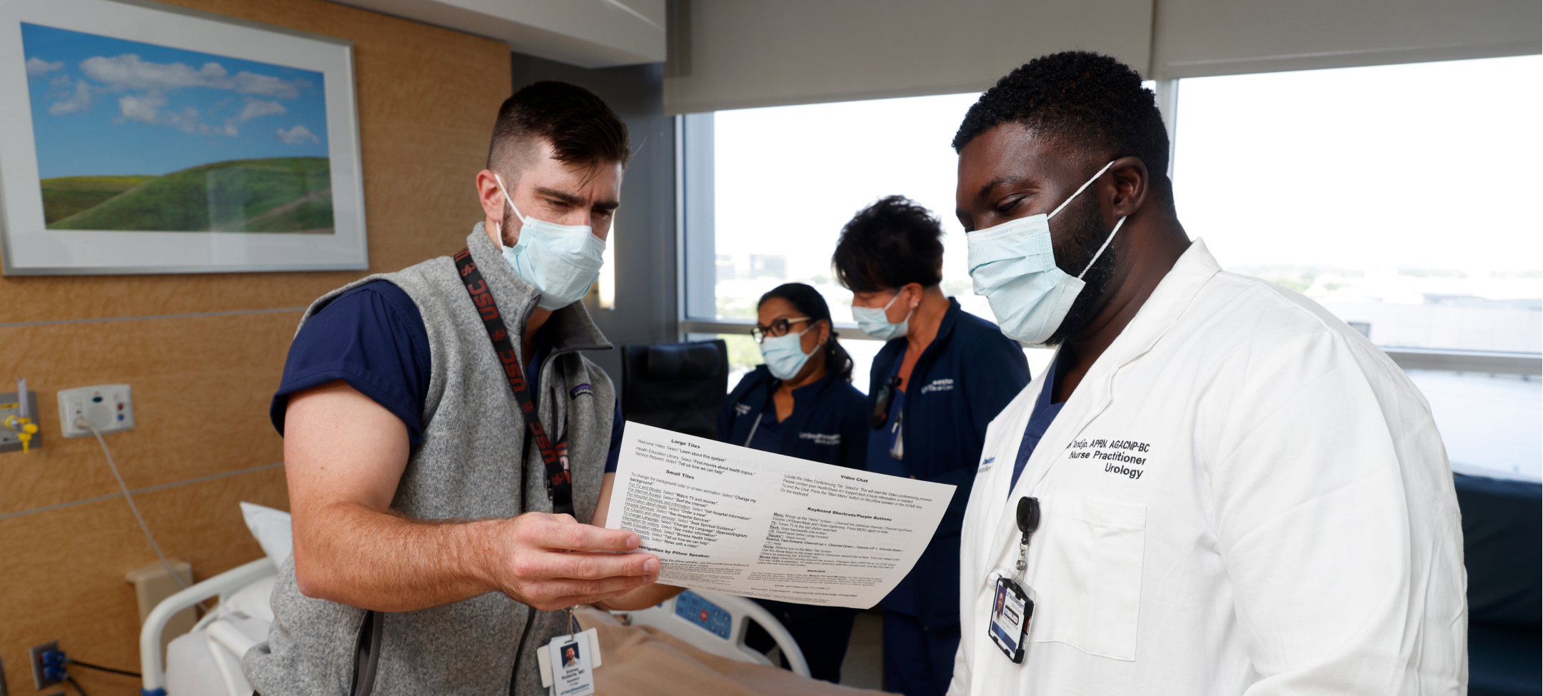 Two male UT Southwestern advanced practice providers (APP’s) looking over a piece of paper. Two healthcare women working in background
