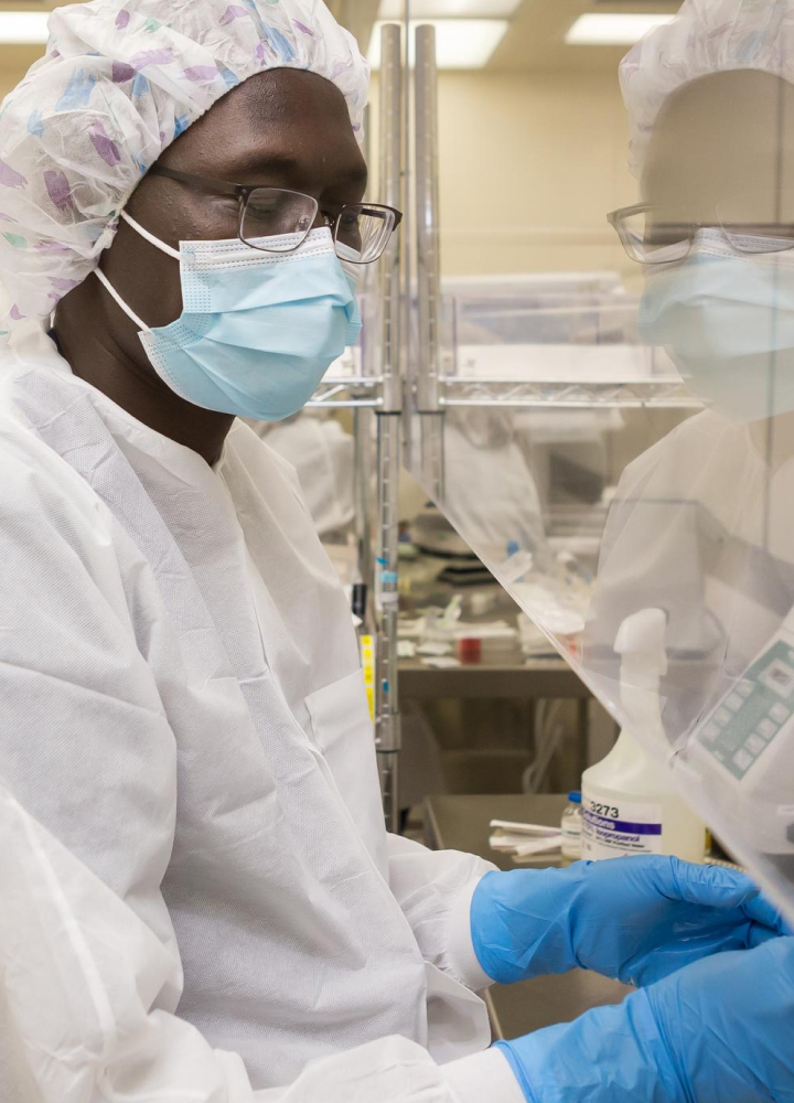 A healthcare worker in full protective gear, including a mask, hair covering, and gloves, works in a sterile environment with medical equipment.