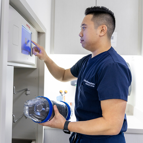 A male healthcare worker in navy scrubs operates a touchscreen panel while holding a medical transport canister in a clinical setting.