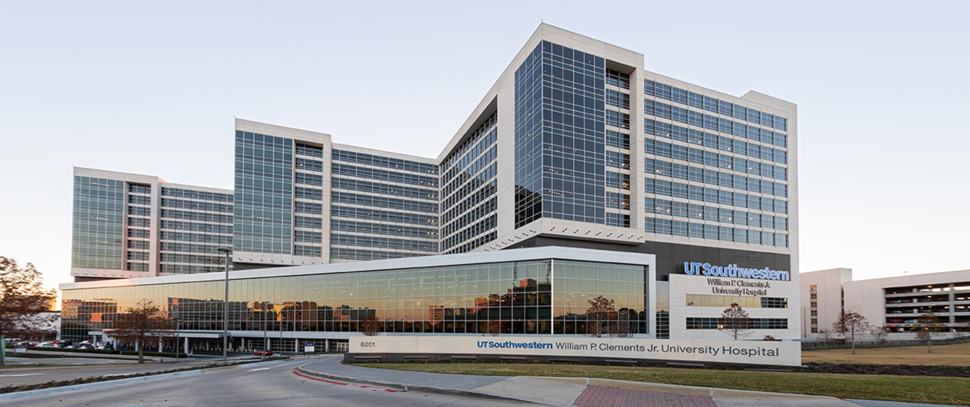 UT Southwestern William P. Clements Jr. University Hospital, a modern glass building reflecting the sunset, with a parking garage nearby.
