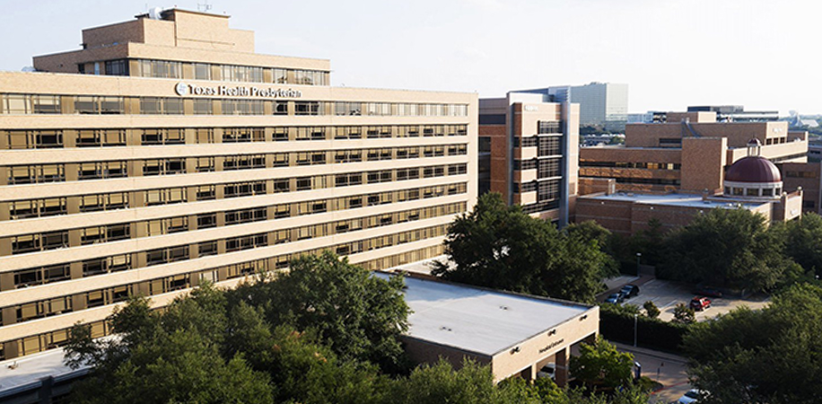 Texas Health Presbyterian Hospital Dallas, a large multi-story medical building surrounded by trees and greenery under a clear sky.