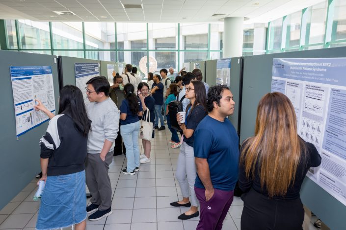 People gather in a bright hallway to view and discuss scientific research posters displayed on partition walls.