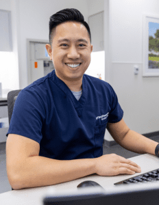 Benefits (3) Male healthcare professional in blue scrubs sitting within a hospital setting and smiling at the camera.