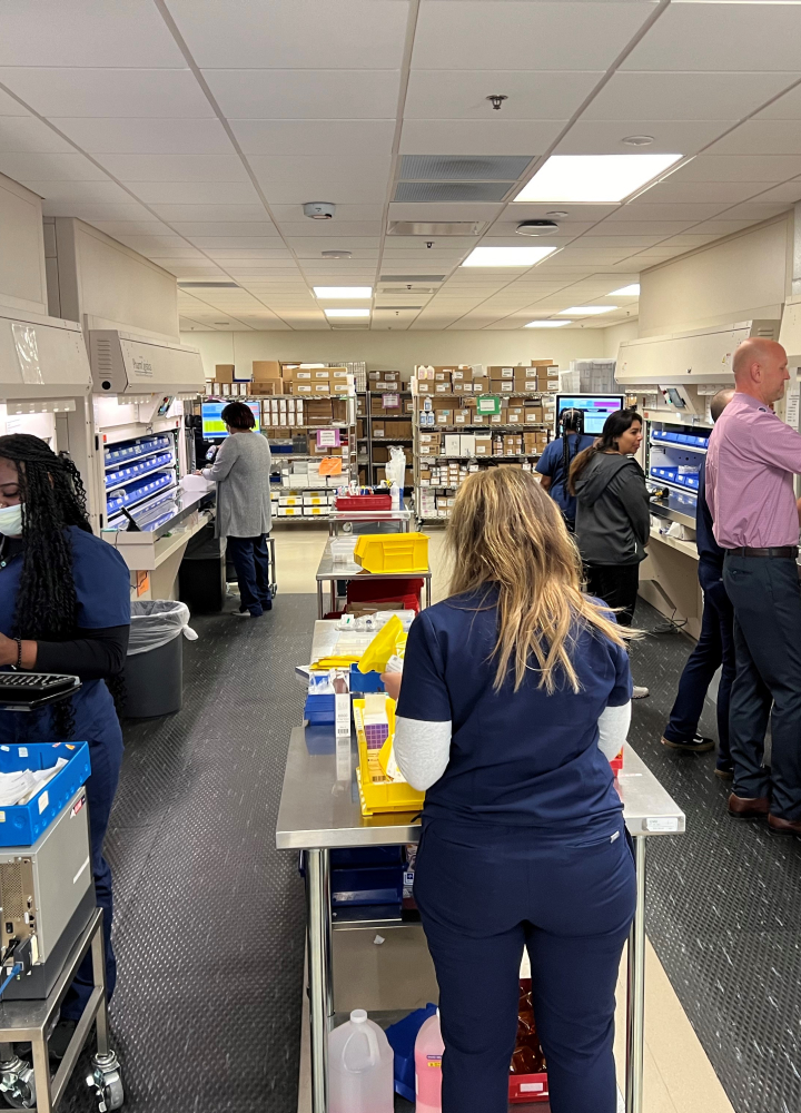 Staff in scrubs work in a busy medical supply room, organizing items on shelves and counters.
