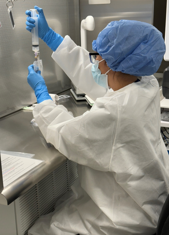 A healthcare worker wearing full protective gear, including a mask, gloves, and hair covering, prepares a syringe in a sterile medical environment.