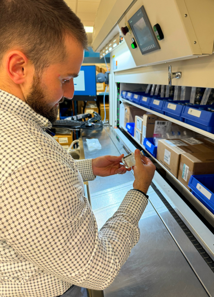 A male employee in a checkered shirt examines a small item in a medical storage area with shelves containing boxes and bins.