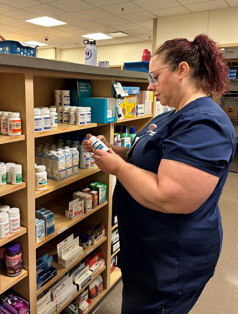 A staff member in scrubs inspects a medication bottle while standing in front of shelves filled with various medications and supplies.
